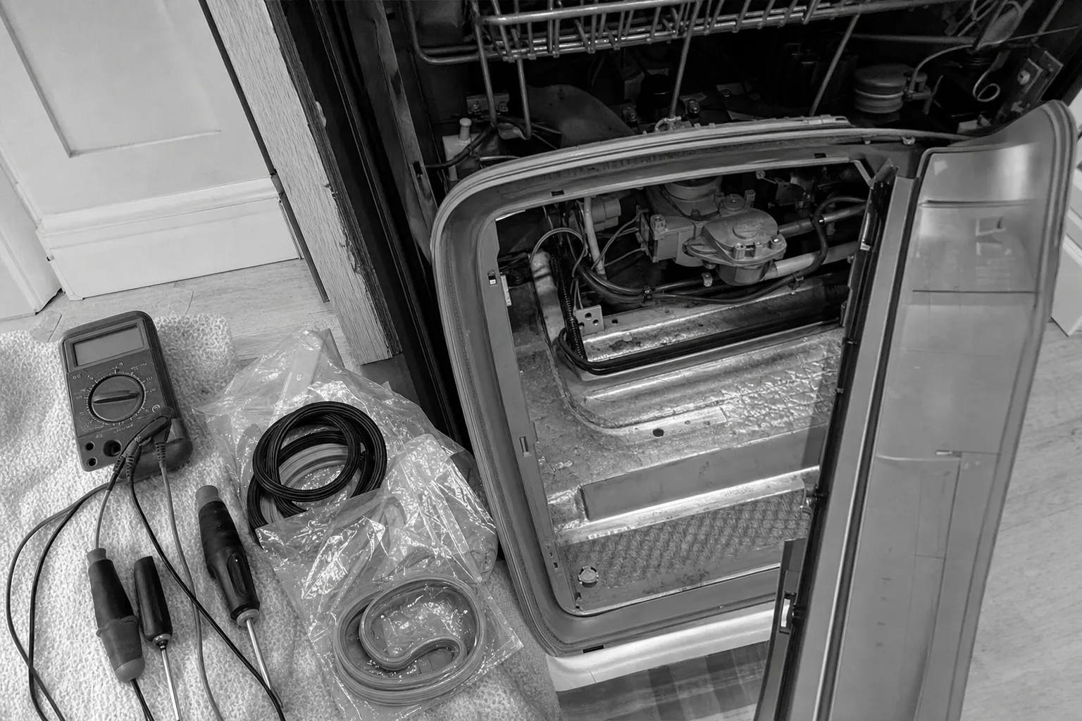 Black-and-white: a side-hinged door swung open on a stripped lower compartment with a motor and wiring exposed, bagged gaskets and a multimeter on a towel