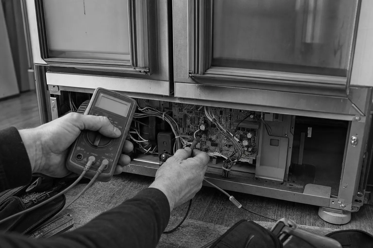 Black-and-white: test leads on a circuit board in the base of a unit with two glass doors, the multimeter held in the other hand